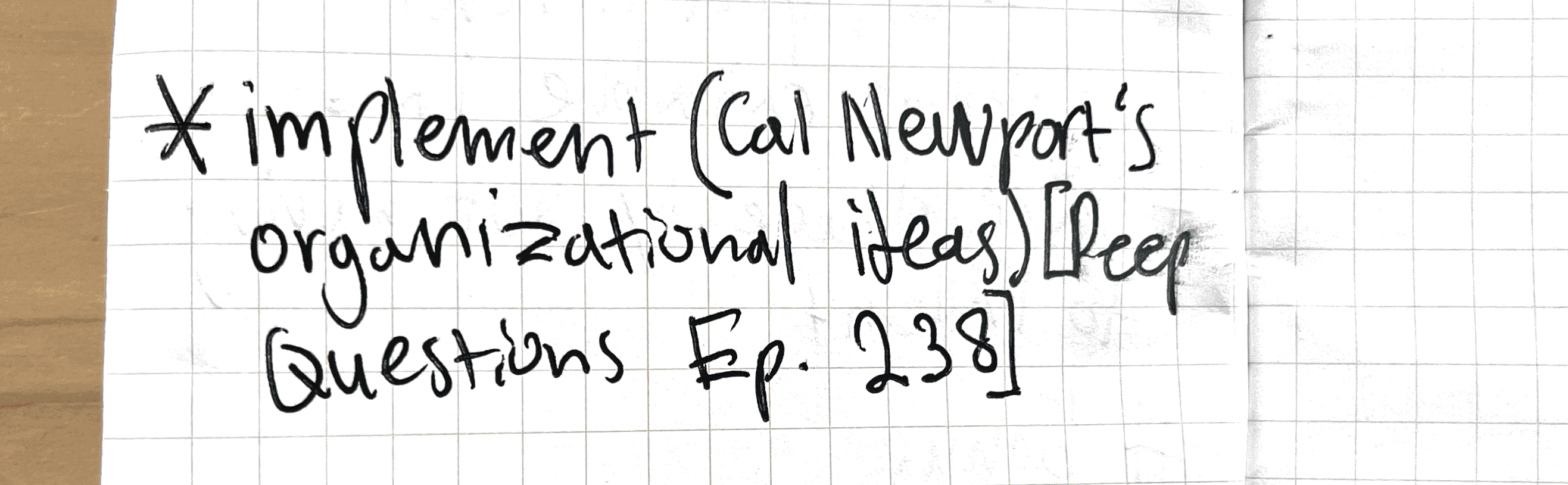 Close-up photo of a handwritten note on grid paper with the text ‘Implement Cal Newport’s organizational ideas)Deep Questions Ep. 238).